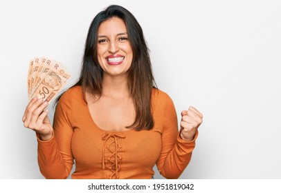 Beautiful hispanic woman holding 50 brazilian real banknotes screaming proud, celebrating victory and success very excited with raised arms 
