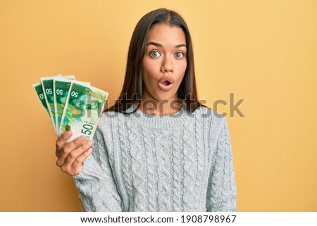 Beautiful hispanic woman holding 50 israel shekels banknotes scared and amazed with open mouth for surprise, disbelief face 