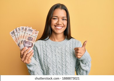 Beautiful hispanic woman holding 500 mexican pesos banknotes smiling happy and positive, thumb up doing excellent and approval sign 