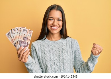 Beautiful hispanic woman holding 500 mexican pesos banknotes screaming proud, celebrating victory and success very excited with raised arm 