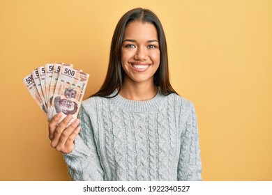 Beautiful hispanic woman holding 500 mexican pesos banknotes looking positive and happy standing and smiling with a confident smile showing teeth 