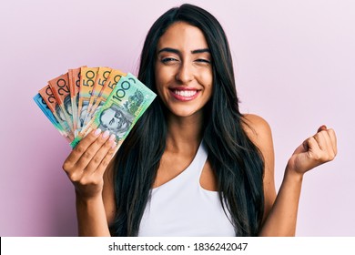 Beautiful hispanic woman holding australian dollars screaming proud, celebrating victory and success very excited with raised arm 