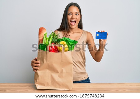 Beautiful hispanic woman holding groceries and credit card celebrating crazy and amazed for success with open eyes screaming excited. 