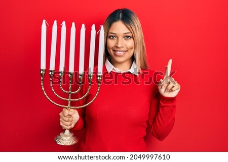 Beautiful hispanic woman holding menorah hanukkah jewish candle smiling with an idea or question pointing finger with happy face, number one 