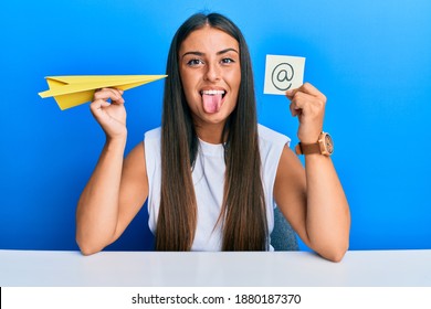 Beautiful Hispanic Woman Holding Paper Plane And Email Symbol Sticking Tongue Out Happy With Funny Expression. 