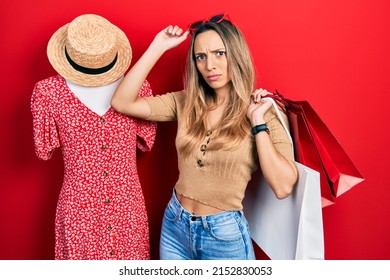 Beautiful Hispanic Woman Holding Shopping Bags By Retail Manikin Clueless And Confused Expression. Doubt Concept. 