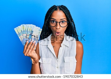Beautiful hispanic woman holding south african 100 rand banknotes scared and amazed with open mouth for surprise, disbelief face 