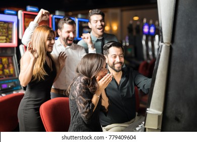 Beautiful Hispanic woman looking excited about hitting the jackpot in a slot machine while her friends celebrate with her