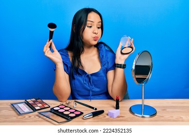 Beautiful Hispanic Woman With Nose Piercing Holding Makeup Powder And Brush Puffing Cheeks With Funny Face. Mouth Inflated With Air, Catching Air. 