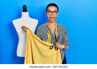 Beautiful hispanic woman with short hair standing by manikin holding cloth afraid and shocked with surprise and amazed expression, fear and excited face. 
