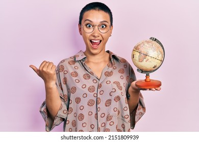Beautiful hispanic woman with short hair holding small world ball pointing thumb up to the side smiling happy with open mouth 