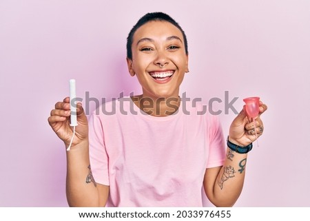 Beautiful hispanic woman with short hair holding menstrual cup and tampon smiling and laughing hard out loud because funny crazy joke. 