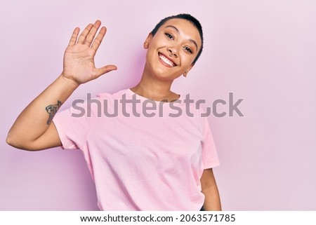 Beautiful hispanic woman with short hair wearing casual pink t shirt waiving saying hello happy and smiling, friendly welcome gesture 