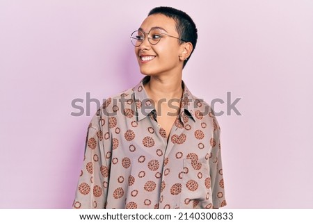 Beautiful hispanic woman with short hair wearing glasses looking away to side with smile on face, natural expression. laughing confident. 
