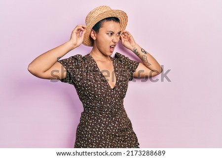 Beautiful hispanic woman with short hair wearing summer hat angry and mad screaming frustrated and furious, shouting with anger. rage and aggressive concept. 