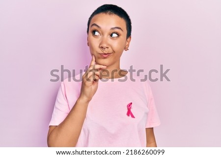 Beautiful hispanic woman with short hair wearing pink cancer ribbon on shirt serious face thinking about question with hand on chin, thoughtful about confusing idea 