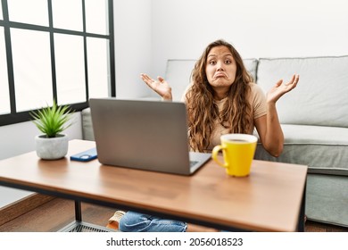 Beautiful Hispanic Woman Using Computer Laptop At Home Clueless And Confused Expression With Arms And Hands Raised. Doubt Concept. 