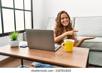Beautiful Hispanic Woman Using Computer Laptop At Home Amazed And Smiling To The Camera While Presenting With Hand And Pointing With Finger. 