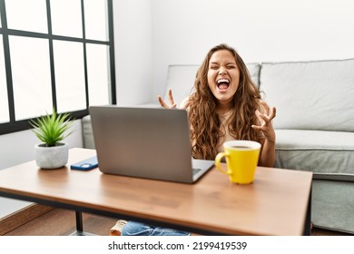 Beautiful Hispanic Woman Using Computer Laptop At Home Crazy And Mad Shouting And Yelling With Aggressive Expression And Arms Raised. Frustration Concept. 