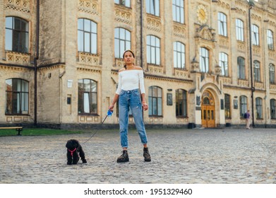 Beautiful Hispanic Woman Walking With A Little Black Dog On A Leash On The Street Against The Backdrop Of A Historic Old Building, Looking At The Camera.