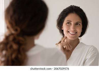 Beautiful Hispanic woman wear white bathrobe admire face view looking in mirror while standing in bathroom enjoy facial perfectness after skin procedure, applied cream, treatment for skincare beauty
