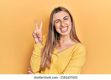 Beautiful hispanic woman wearing casual yellow sweater smiling with happy face winking at the camera doing victory sign with fingers. number two. 