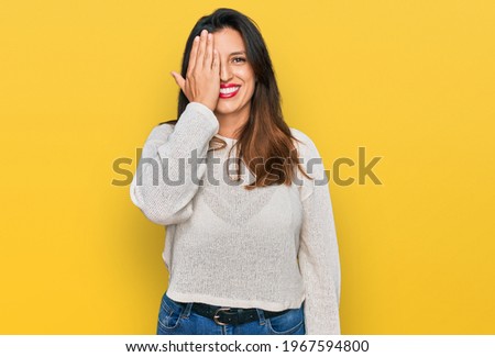 Beautiful hispanic woman wearing casual sweater covering one eye with hand, confident smile on face and surprise emotion. 