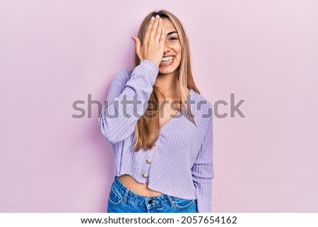 Beautiful hispanic woman wearing casual shirt covering one eye with hand, confident smile on face and surprise emotion. 