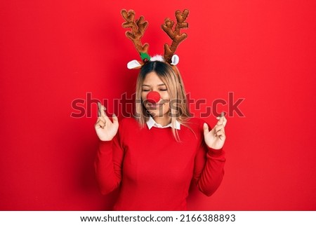 Beautiful hispanic woman wearing deer christmas hat and red nose gesturing finger crossed smiling with hope and eyes closed. luck and superstitious concept. 