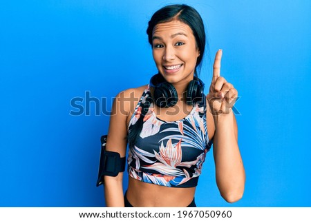 Beautiful hispanic woman wearing gym clothes and using headphones smiling with an idea or question pointing finger up with happy face, number one 