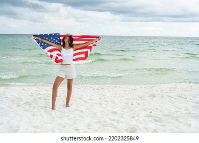  Beautiful Hispanic woman in a white bikini with an American flag on whet sandy beach emerald waters of the emerald coast the Gulf Of Mexico with a hat and a drink and beach bag  - Powered by Shutterstock