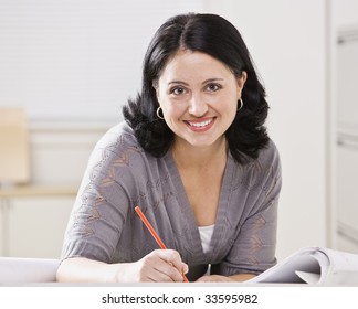 Eine schöne hispanische Frau, die an einem Schreibtisch schreibt.  Sie lächelt die Kamera an.  Viereckige Komposition.