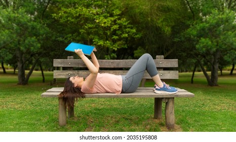 Beautiful Hispanic young woman in sportswear lying on the wooden bench in the middle of a park without people reading a blue book against a background of green trees