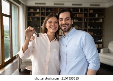 Beautiful Homeowners Couple, 30s Wife And Husband Hugging Pose In Fashionable Living Room Holding Keys Bunch Feeling Happy At Relocation Day. Bank Loan, Real-estate Buyers, First Home, Tenancy Concept