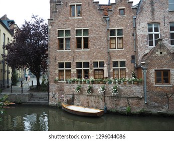 A Beautiful House On The Water With Pots Of Flowers And A Wooden White Boat Next To Autumn Trees And An Older Couple Walking Hand In Hand In The Background