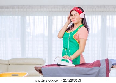 Beautiful Housewife Enjoying A Music On Headphones While Ironing Clothes In The Living Room At Home