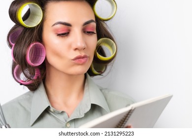 Beautiful Housewife. Young Cheerful Woman With Hair Curlers, Bright Make-up, A Notebook And A Whisk In Her Hands, On A White Background. Thinking About The Recipe For Dinner.