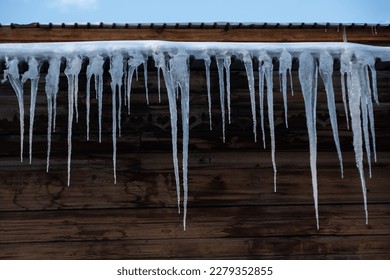 Beautiful huge icicles hang from the roof of the building. Melting icicles in the rays of the spring sun. Ice dam in gutter and ice frozen on roof in winter. Icicles falling danger concept.
