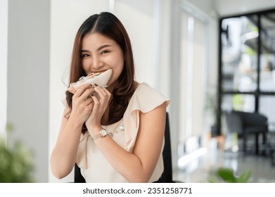 A beautiful, hungry Asian businesswoman is having breakfast, eating a sandwich at her desk in the office. - Powered by Shutterstock