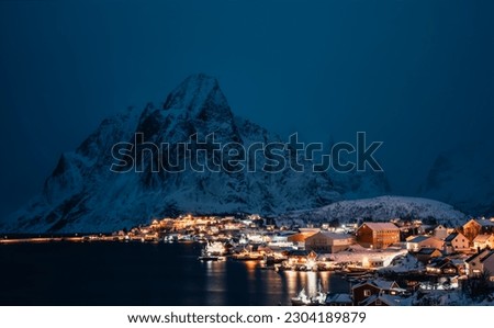 A beautiful illuminated building stands against a majestic mountain range, blanketed in snow and bathed in the night sky.