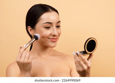 Beautiful Indian Female Holding Compact Powder And Applying On Face With Brush, Smiling Young Hindu Woman Looking In Mirror While Doing Daily Makeup Over Beige Studio Background, Closeup Shot