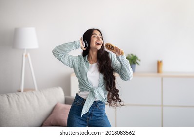 Beautiful Indian lady with headphones dancing and singing, using hairbrush as microphone at home. Millennial woman moving to her favorite song, enjoying music, pretending to be popular star
