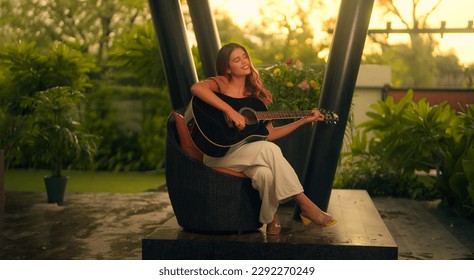 Beautiful Indian woman sitting on chair playing acoustic guitar and singing alone outside home. Happy Cheerful female enjoy sing some new sound on classical guitar Learning, practicing as hobby  - Powered by Shutterstock