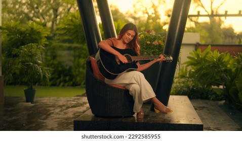 Beautiful Indian woman sitting on chair playing acoustic guitar and singing alone outside home. Happy Cheerful female enjoy sing some new sound on classical guitar Learning, practicing as hobby - Powered by Shutterstock