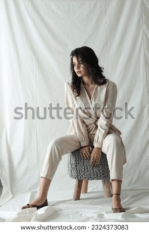 Beautiful Indian woman sitting on a puffy table natural makeup wear fashion clothes casual dress code office style total cream pants suit, romantic date business meeting accessory armchair interior