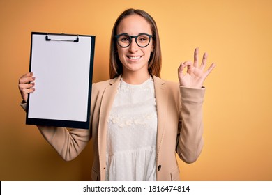 Beautiful Inspector Woman Wearing Glasses Holding Checklist Clipboard Over Yellow Background Doing Ok Sign With Fingers, Excellent Symbol