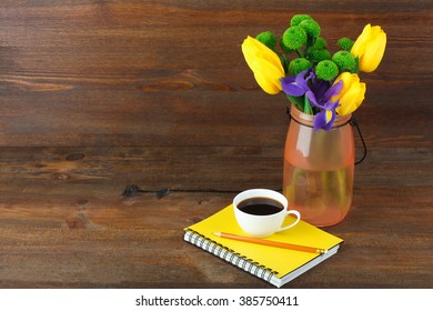 beautiful iris with yellow tulips and green chrysanthemums in a vase near a Notepad with coffee Cup on dark background brown wooden table