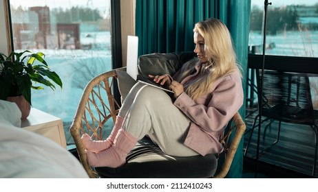 Beautiful Isolated Caucasian Female Sitting On A Comfortable Chair By The Window Using Her Laptop Device To Work From Home, Wearing Cute Grey Sweatpants And A Pink Hoodie. Pandemic Remote Working.