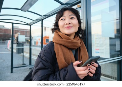 Beautiful Japanese girl, student standing on bus stop in winter jacket, holding smartphone, using application to track her public transport, ordering a ride. - Powered by Shutterstock