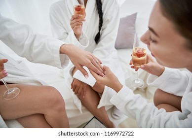 Beautiful jewelry. Ladies in white bathrobes holding glasses of champagne and looking at arm of future bride. Women sitting on daybeds at spa salon